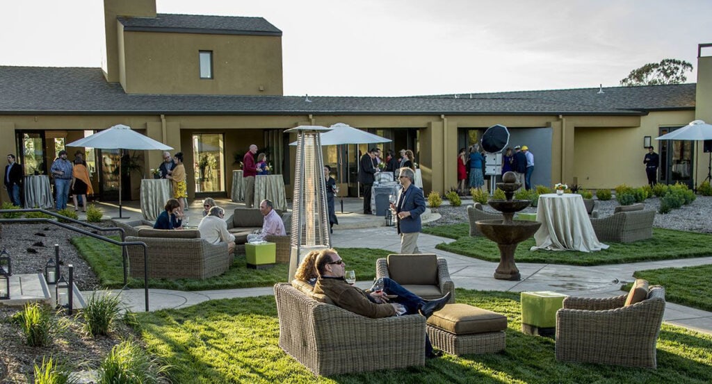 People gather at an outdoor event with wicker seating on green lawns, umbrellas, and tall patio heaters. Some individuals chat near round, cloth-covered tables while others relax on couches. A fountain is in the middle, and the building behind has large windows and earth-toned walls.
