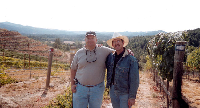 Two men are standing side by side in an outdoor vineyard with rolling hills in the background. The man on the left is wearing a beige polo shirt, blue jeans, and a cap, while the man on the right is wearing a denim jacket, white cowboy hat, and glasses. Both are smiling at the camera.
