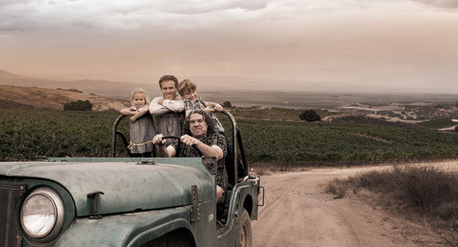 A sepia-toned image of an older man and three children in an open-top jeep on a dirt road near a vast vineyard. The man is seated behind the wheel, and two girls and a boy are standing behind him, smiling. Rolling hills and a cloudy sky span the background.
