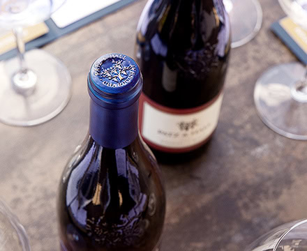 Close-up image of two wine bottles on a table, one with a visible blue foil-wrapped top, bearing a decorative seal. There are multiple wine glasses around the bottles, reflecting ambient light. The background surface appears wooden, with a slightly blurred effect, emphasizing the bottles.
