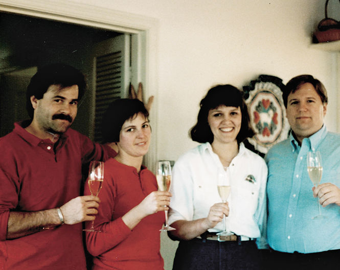Four people are standing indoors, holding champagne glasses and smiling at the camera. They are in a well-lit space with a decorative plate on the wall in the background. The individuals are casually dressed. Two are wearing red tops and two are wearing white tops with one person also in blue.
