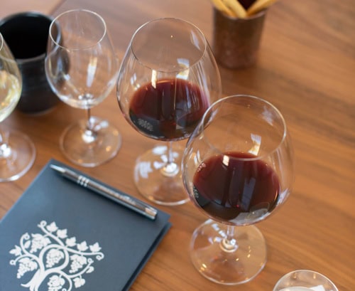 Two glasses filled with red wine are on a wooden table, surrounded by emptied wine glasses. A dark blue notebook with white decorative patterns and an elegant pen rests nearby. In the background, a gold cup holding breadsticks can be seen.
