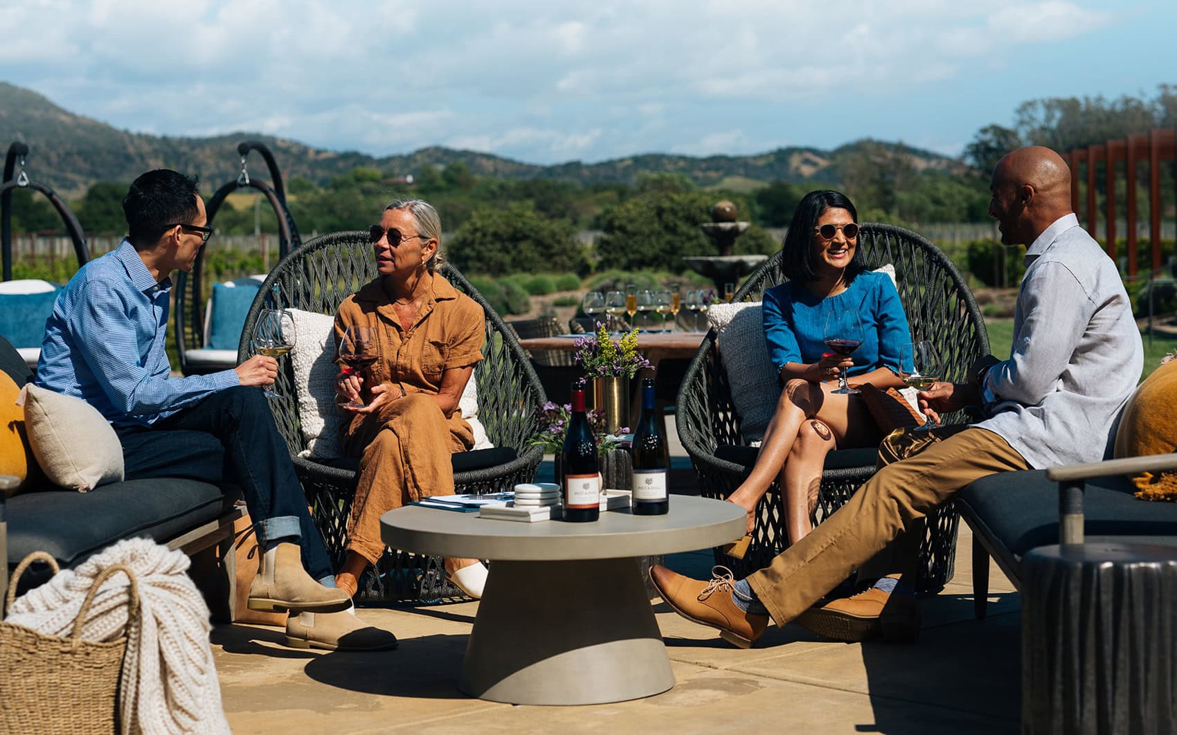 Four individuals sit outdoors at a round table adorned with wine bottles and glasses. They are engaged in conversation and enjoying drinks. The background features green hills and a partly cloudy sky. They are seated on comfortable, modern patio furniture, suggesting a relaxed and social atmosphere.