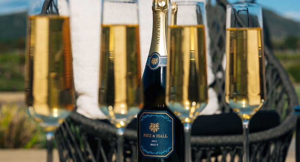 A 2013 bottle of Patz & Hall Brut sparkling wine is displayed prominently, surrounded by four filled champagne flutes. The setting appears to be outdoors, with a blurred background including a wicker couch and greenery. The scene evokes a celebratory and relaxed atmosphere.
