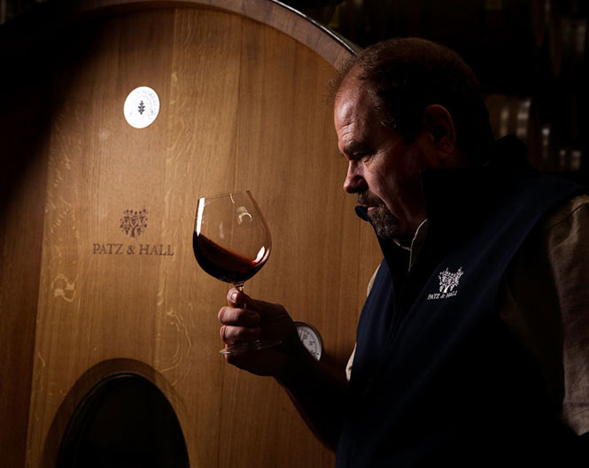 A man with light brown hair and a beard examines a glass of red wine against the dimly lit background of a wooden wine barrel. He is wearing a dark blue vest and a shirt. The barrel features the 