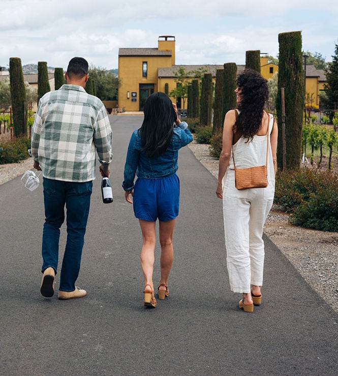 Three people walking down a paved pathway at a vineyard. The person on the left is carrying two wine bottles and wearing a checkered shirt. The person in the middle has a blue romper and is holding a phone. The person on the right wears white overalls and carries a tan purse. Tall trees line the path.
