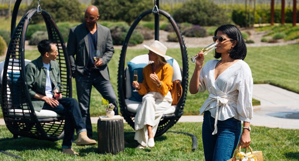 Four people enjoy a sunny day outdoors with drinks. A woman in the foreground sips from a champagne flute and holds a straw purse. Behind her, a man stands with a drink, and another man and woman, seated in hanging chairs, converse. The background features a lawn and plants.
