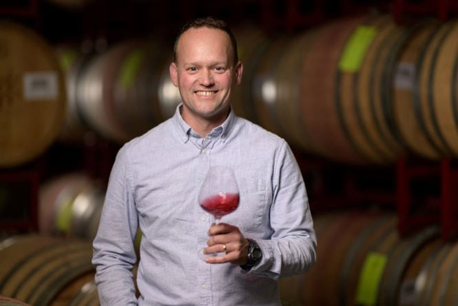 A man with short hair, wearing a light gray button-up shirt, smiles while holding a glass of red wine. He stands in a wine cellar with wooden barrels stacked in the background. The ambiance is warm and inviting, highlighting the wine barrels behind him.
