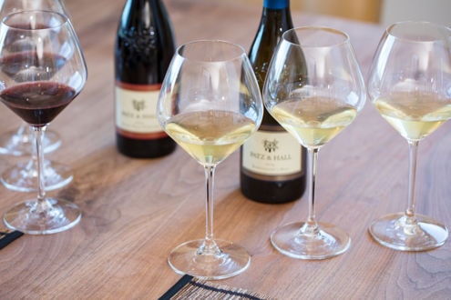 Three wine glasses filled with white wine and one glass with red wine are arranged on a wooden table. In the background, there are three wine bottles, each labeled 