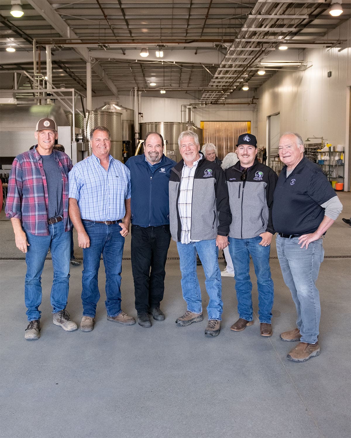 Six men stand side by side inside a spacious industrial building with high ceilings and bright lighting. They are wearing casual and semi-casual clothing, including plaid shirts, jackets, and jeans. The background shows industrial equipment and a person in the distance. Everyone is smiling.
