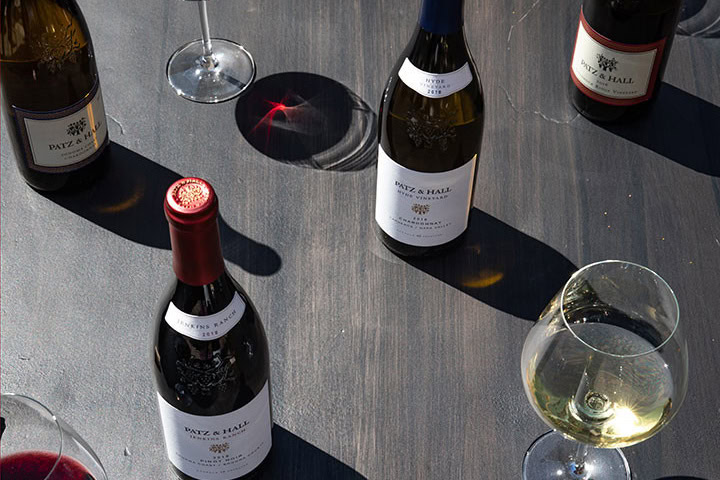 Four bottles of Patz & Hall wine and three wine glasses are placed on a dark wooden table. Two glasses contain white wine, one glass contains red wine. The sunlight casts shadows and a red reflection on the table. The wines appear to have labels with detailed black text and logo.
