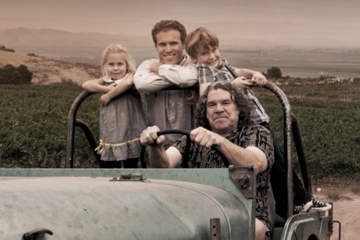A smiling family poses on a green vintage tractor in a rustic, rural setting. The elderly man sits in the driver's seat, while a young girl stands on the left and a boy leans on the right, both resting on the arms of a man standing behind them. Rolling hills and farmland are visible in the background.
